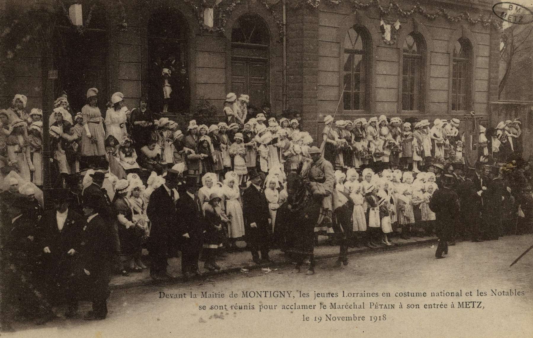 Contenu du Devant la Mairie de MONTIGNY, les jeunes lorraines en costume national et les Notables se sont réunis pour acclamer le Maréchal Pétain à son arrivée à Metz, le 19 Novembre 1918
